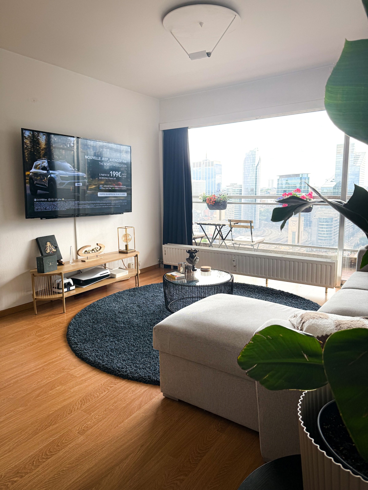 A spacious living room features a large, comfortable sofa arranged around a round rug. A flat-screen television is mounted on the wall, and a small table with two chairs offers a view of the city through expansive windows. Potted plants add a touch of greenery.