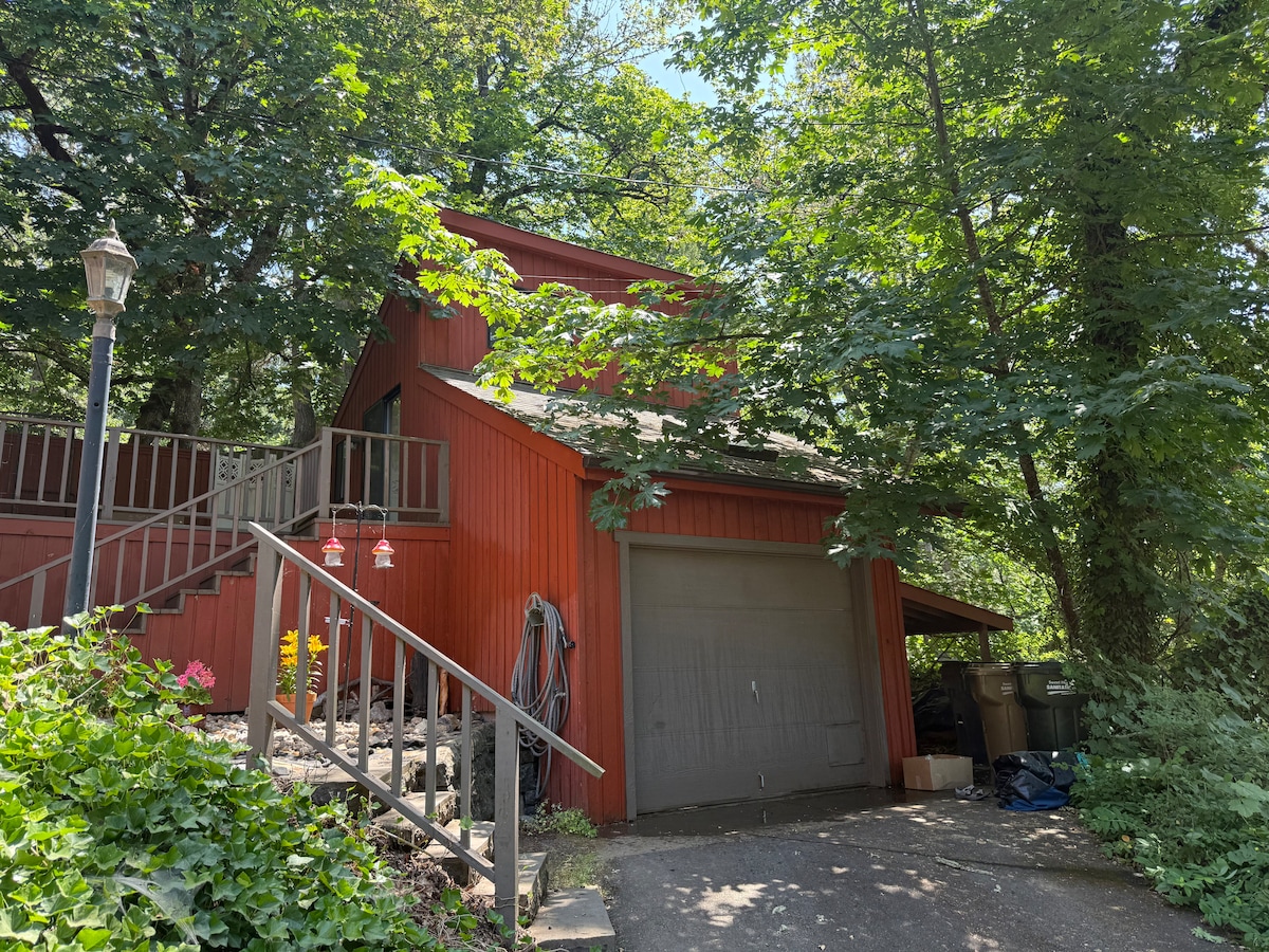 A charming red house is nestled among lush greenery, with a sloped driveway leading up to the entrance. Sturdy wooden steps provide access to a porch. The garage door is visible and surrounded by leafy trees, creating a serene and private atmosphere.