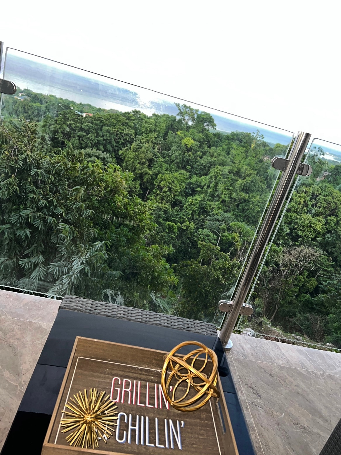 A balcony railing made of glass offers an unobstructed view of the lush, green landscape below. A wooden tray displays the words 'GRILLIN CHILLIN' accompanied by a decorative orb, adding a touch of style to the outdoor space.