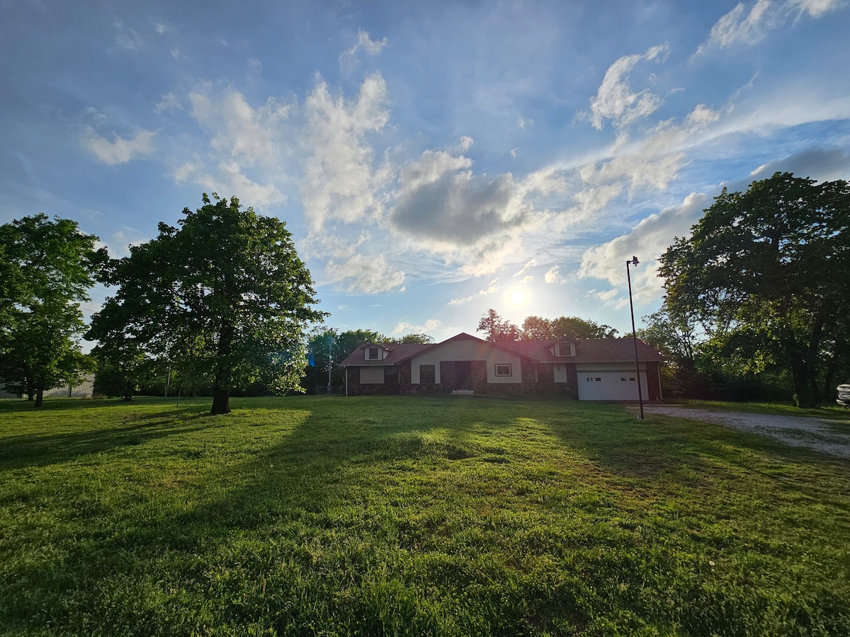 The exterior of the home is presented under a bright sky, with sunlight filtering through scattered clouds. Lush green grass surrounds the house, which features a mix of brick and siding. Tall trees are positioned on either side, casting gentle shadows on the lawn.