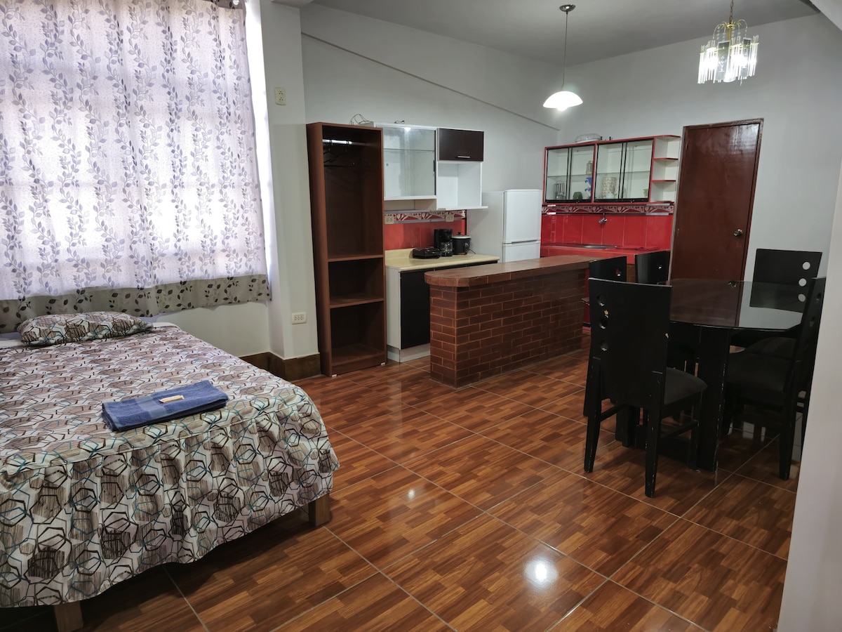 An open living space features a spacious bed with patterned bedding, a dining area with a round table and four chairs, and a red-equipped kitchen counter in the background. Natural light enters through a large window with sheer curtains, enhancing the warm atmosphere.
