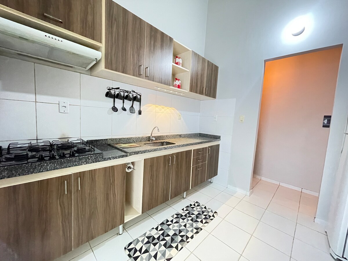A modern kitchen featuring wood cabinetry with open shelving highlights. A gas stove is installed against a tiled backsplash, and a sink is positioned under a window. A patterned rug adds contrast to the tiled floor, while soft lighting illuminates the space.
