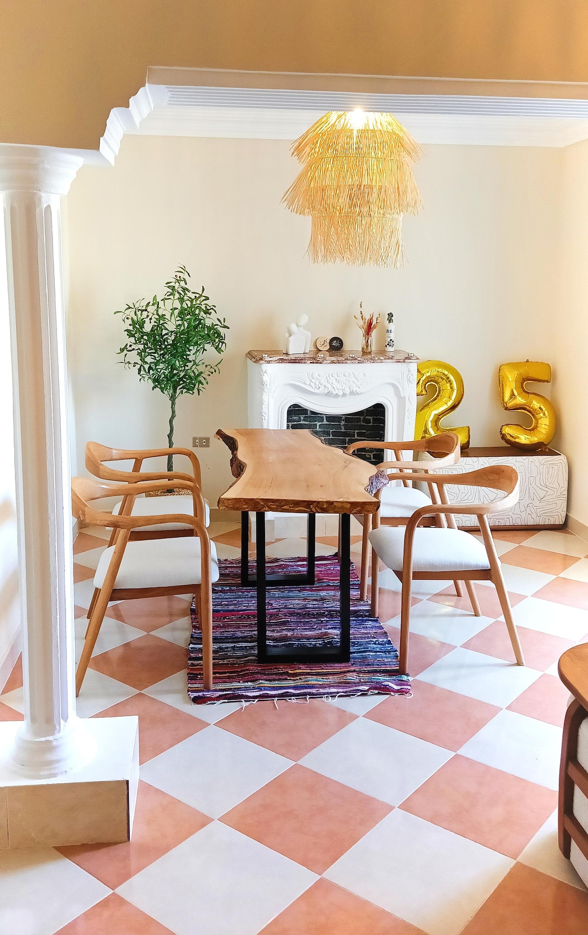 A dining area features a wooden table surrounded by three wooden chairs and one upholstered chair. A decorative rug is placed beneath the table. A potted plant adds a touch of greenery, while a fireplace is decorated with figurines. Gold balloons indicate a celebration.