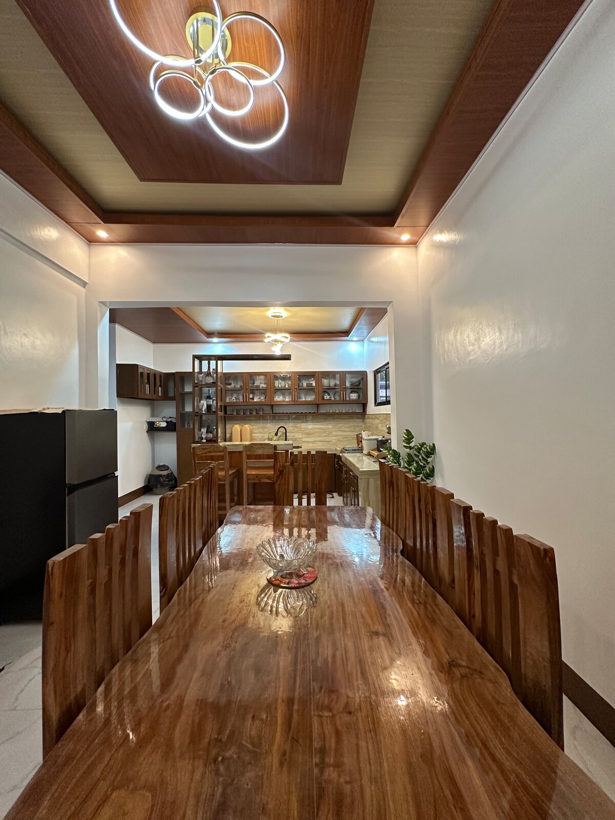 A spacious dining area features a long wooden table surrounded by tall wooden chairs. A modern light fixture hangs from the ceiling, illuminating the room. The kitchen area in the background is equipped with dark cabinetry and a countertop, providing a functional cooking space.