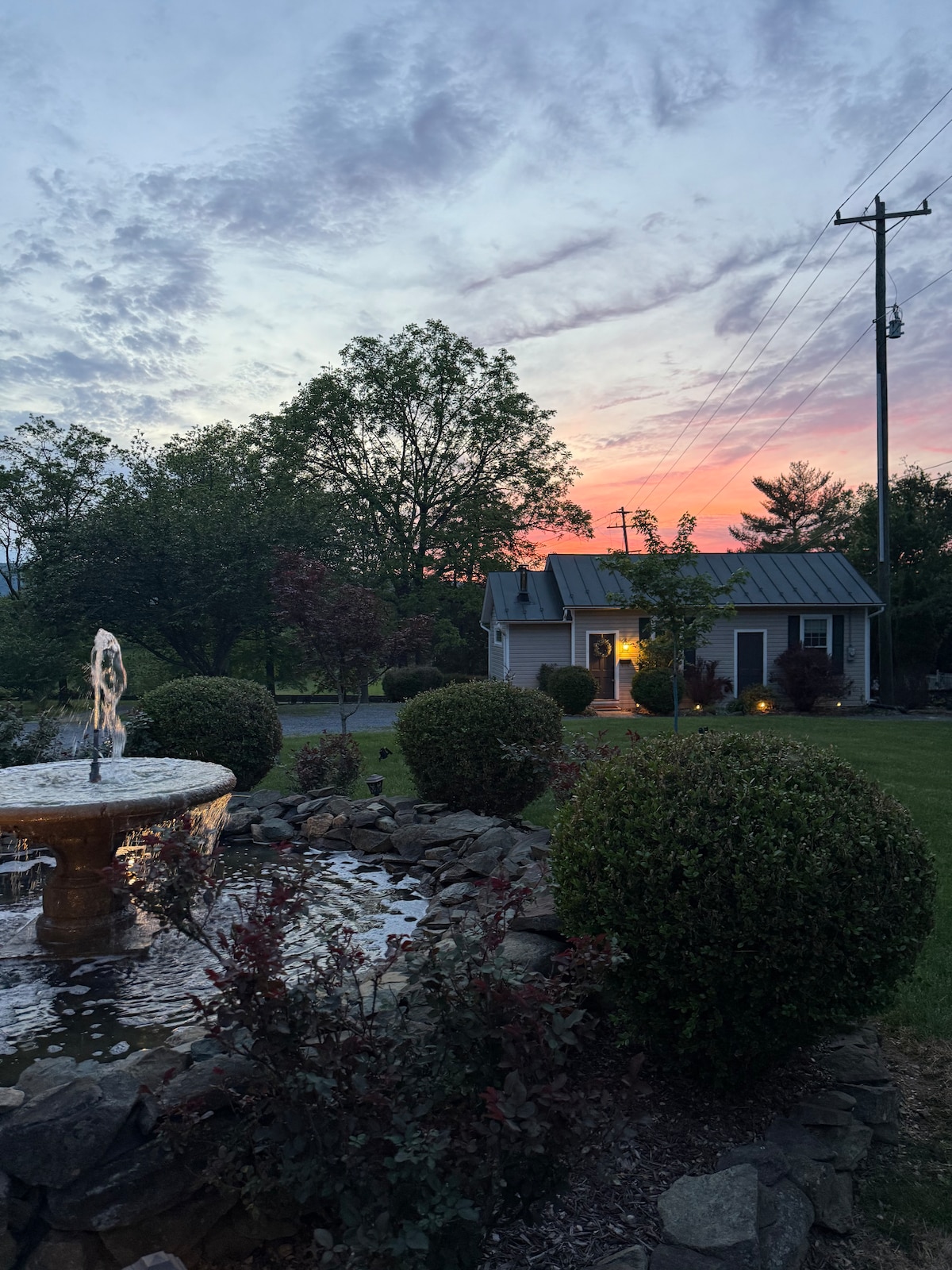 A charming cottage is illuminated by warm light, set against a vibrant sunset sky. A serene fountain with flowing water is featured in the foreground, surrounded by neatly trimmed shrubs and landscaped lawn, enhancing the peaceful ambiance of the outdoor space.