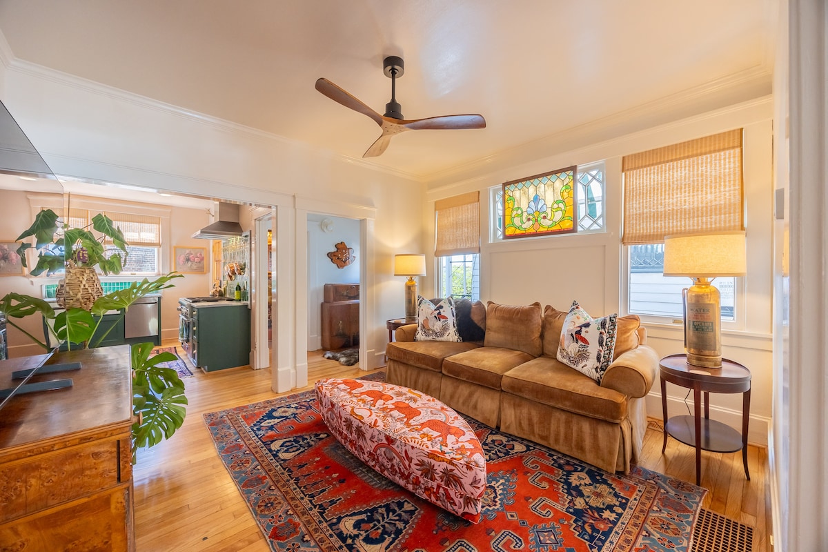A welcoming living area features a plush brown sofa with decorative pillows, complemented by a vibrant orange patterned ottoman. Natural light filters through stained glass windows, while woven shades provide privacy. A colorful Persian rug adds warmth to the hardwood floor, enhancing the room's inviting ambiance.