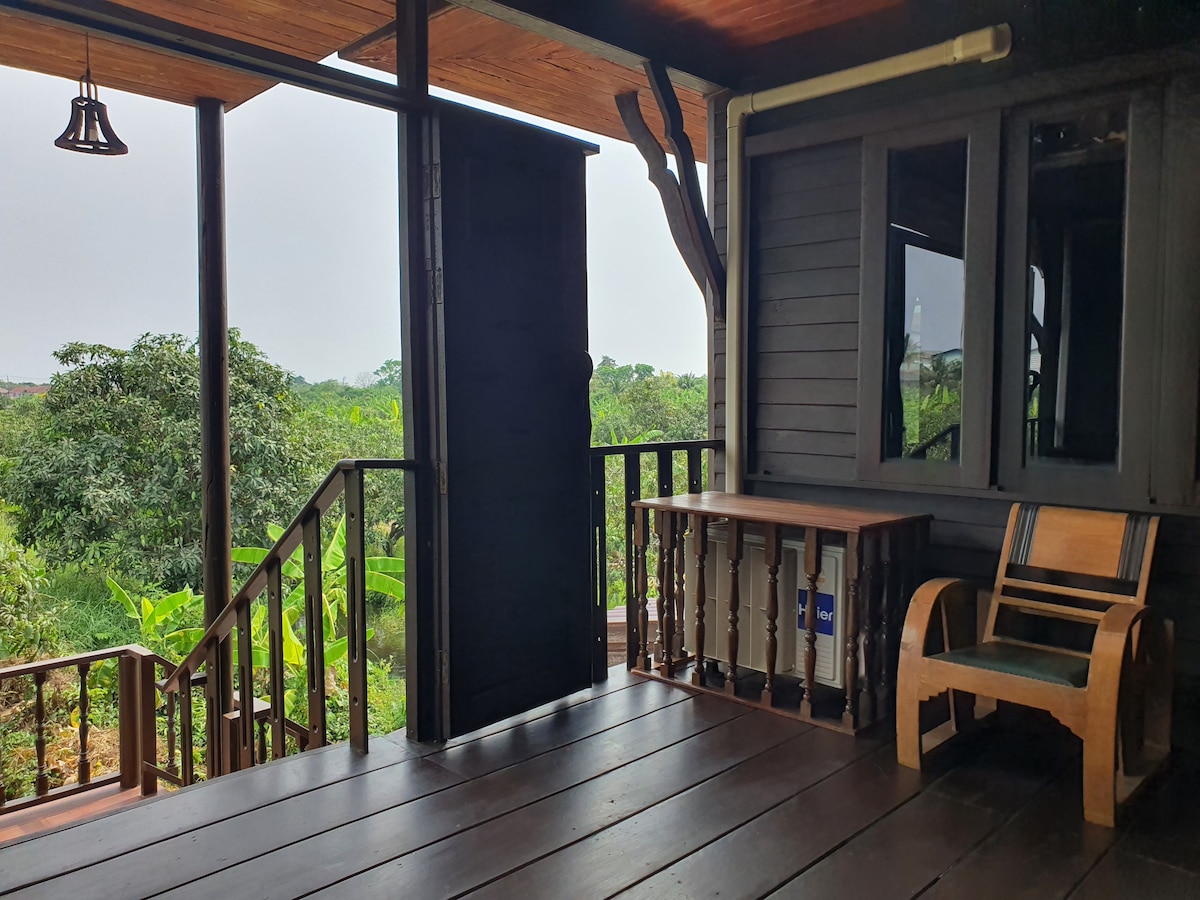 An outdoor space features wooden flooring with a railing overlooking a lush landscape of greenery. A simple wooden table and an armchair provide seating. Behind, a single door leads indoors, while a bell adds a charming touch to the peaceful setting.