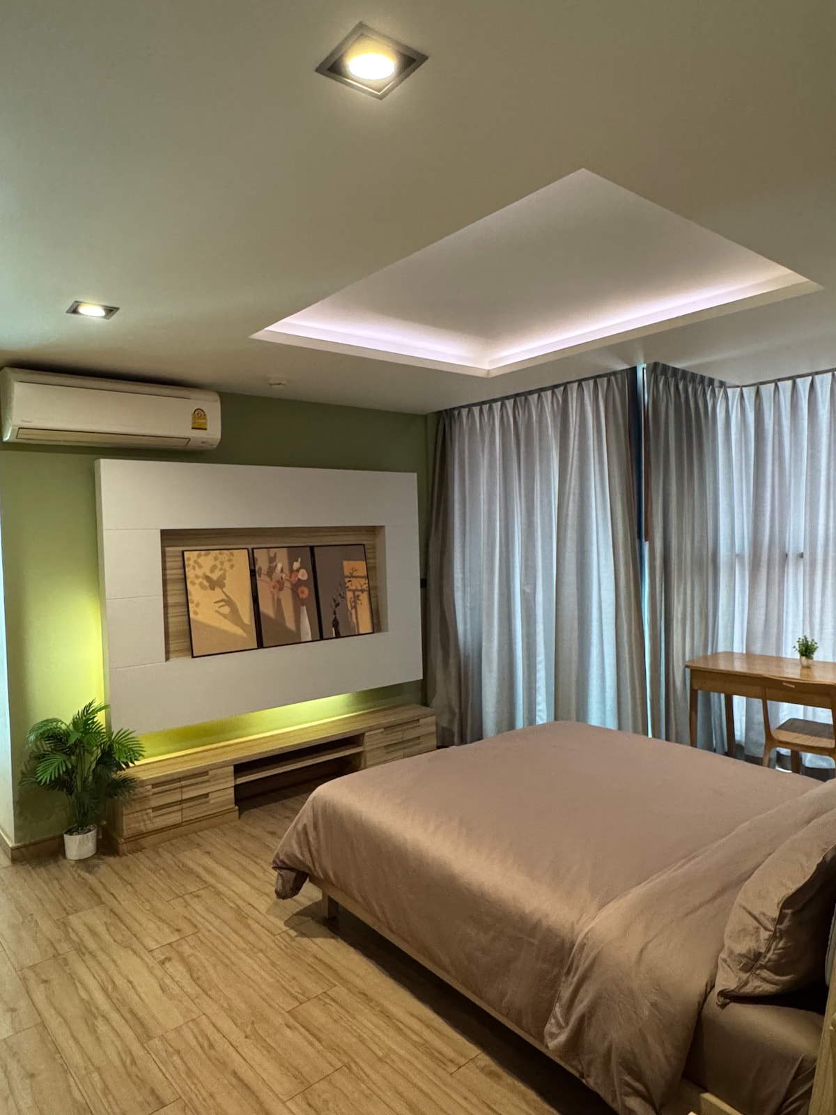 A spacious bedroom is equipped with a comfortable bed and soft bedding. Large windows are dressed with sheer curtains, allowing natural light to illuminate the room. A small dining area is included, complemented by a decorative plant and modern wall art.