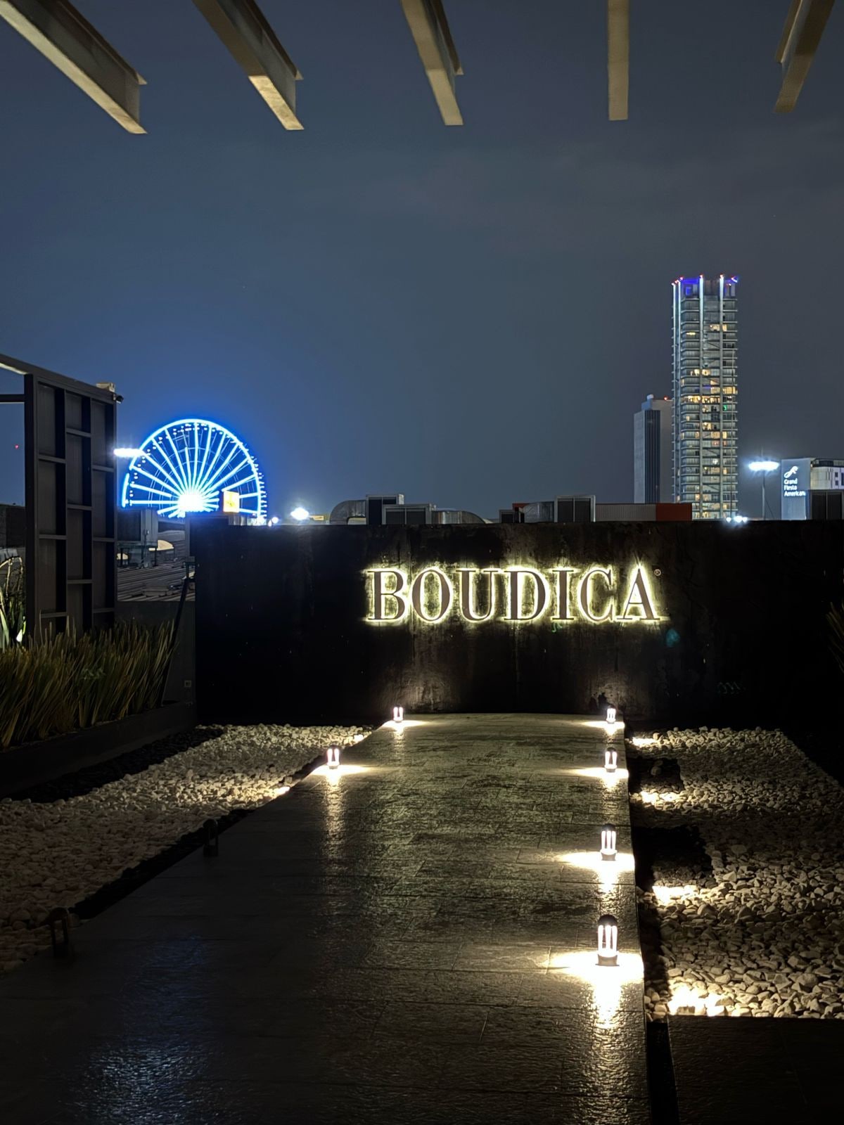 The entrance to Boudica is marked by illuminated signage, with soft lighting casting a warm glow on the pathway. Pebbles line the edges of the walkway, which leads towards a backdrop of a brightly lit Ferris wheel and city skyline at night.