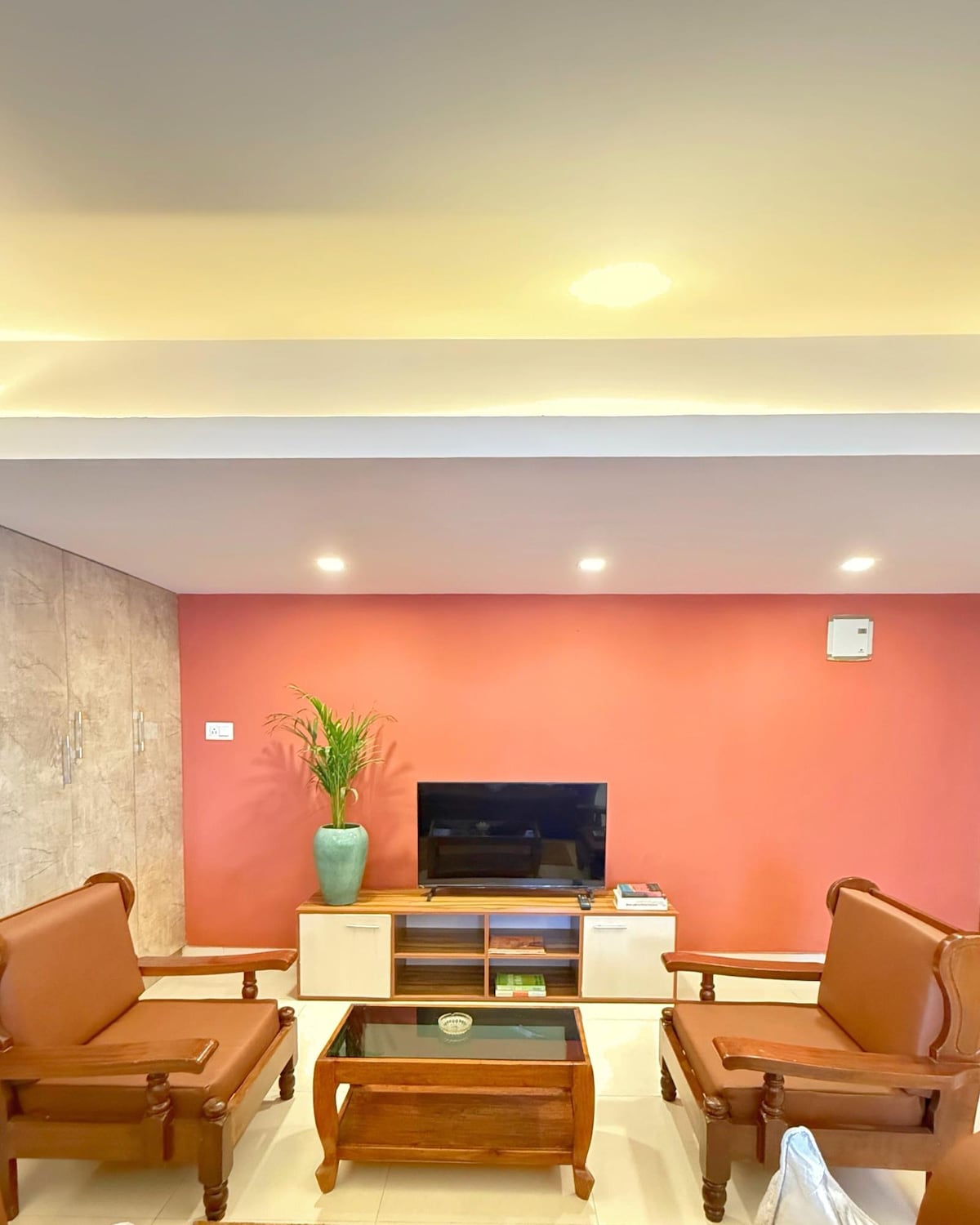 A comfortable living area is presented with two brown armchairs facing a sleek coffee table. A television is mounted on the wall against a warm pink backdrop, while a potted plant adds a touch of greenery. Soft lighting enhances the inviting space.