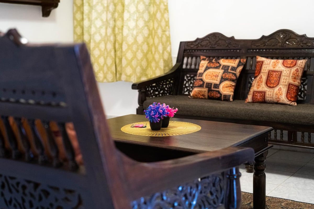 An elegantly crafted wooden sofa is positioned against a light-colored wall, adorned with patterned cushions. A round table is placed in the center, holding a small flower pot. A decorative curtain in soft hues enhances the serene ambiance of the space.