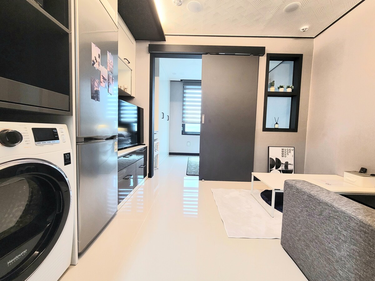 A compact living area features an open layout with a modern design. A washing machine is visible, alongside a refrigerator and a television. Light-colored walls enhance the bright atmosphere, and sleek furniture complements the clean lines of the space.
