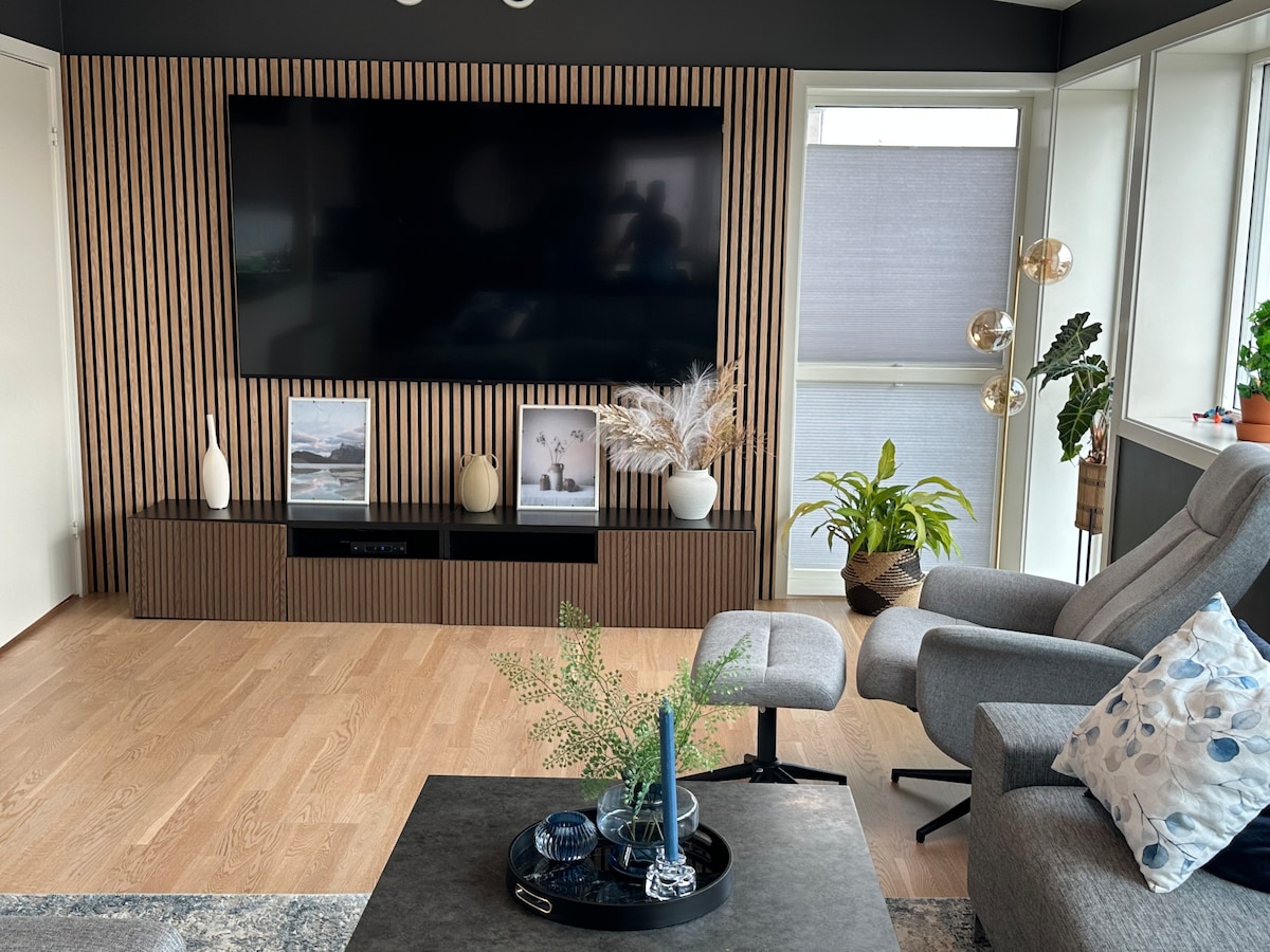A modern living area features a large flat-screen television mounted on a slatted wood wall. A comfortable gray armchair and a coffee table adorned with decorative items are positioned in front. Plants add a touch of greenery, while large windows bring in natural light.