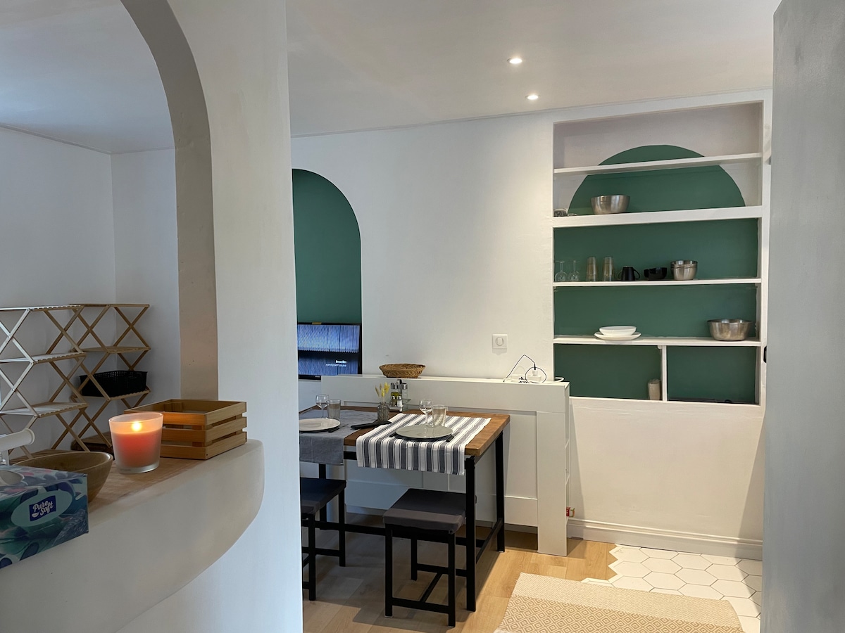 A modern kitchen is displayed, featuring a table with four stools and a striped table runner. Open shelves showcase various dishware against a green wall, while a warm candle adds a soft glow. The space is brightened by recessed lighting and a large archway.