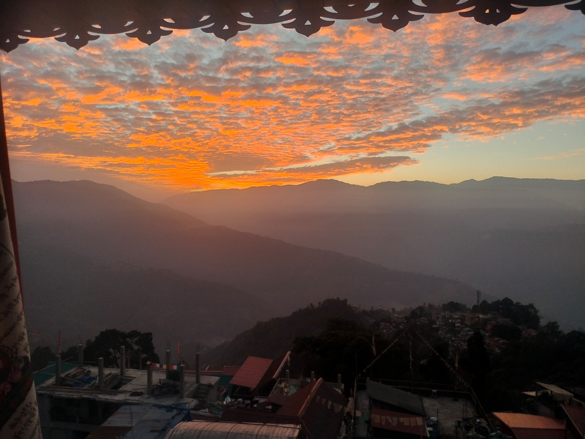 A sunset view is captured, showcasing layers of mountains beneath a colorful sky with clouds in shades of orange and pink. The landscape features a gradual gradient of light to dark, highlighting the horizon and creating a serene atmosphere.