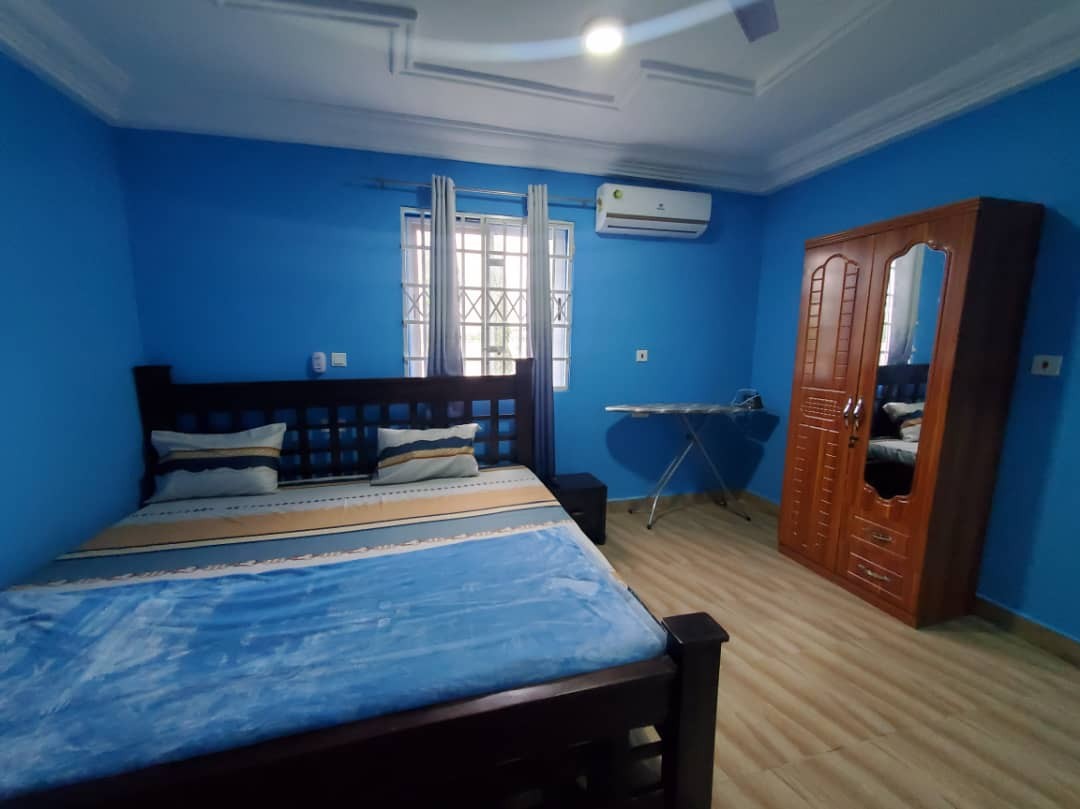 A spacious bedroom is presented with blue walls and a large bed adorned with soft bedding. Natural light enters through a window, complemented by light-colored curtains. An air conditioning unit is mounted on the wall, and a wooden wardrobe stands nearby, adding functionality to the space.