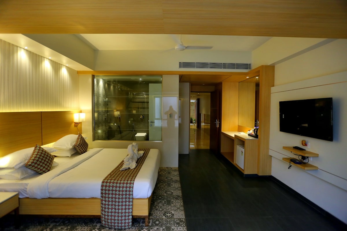 A modern bedroom features a spacious bed with decorative cushions and a neatly arranged towel art figure. The room is illuminated by soft lighting, with a glass-enclosed shower visible in the background. A television and a coffee station are positioned against a wooden wall.