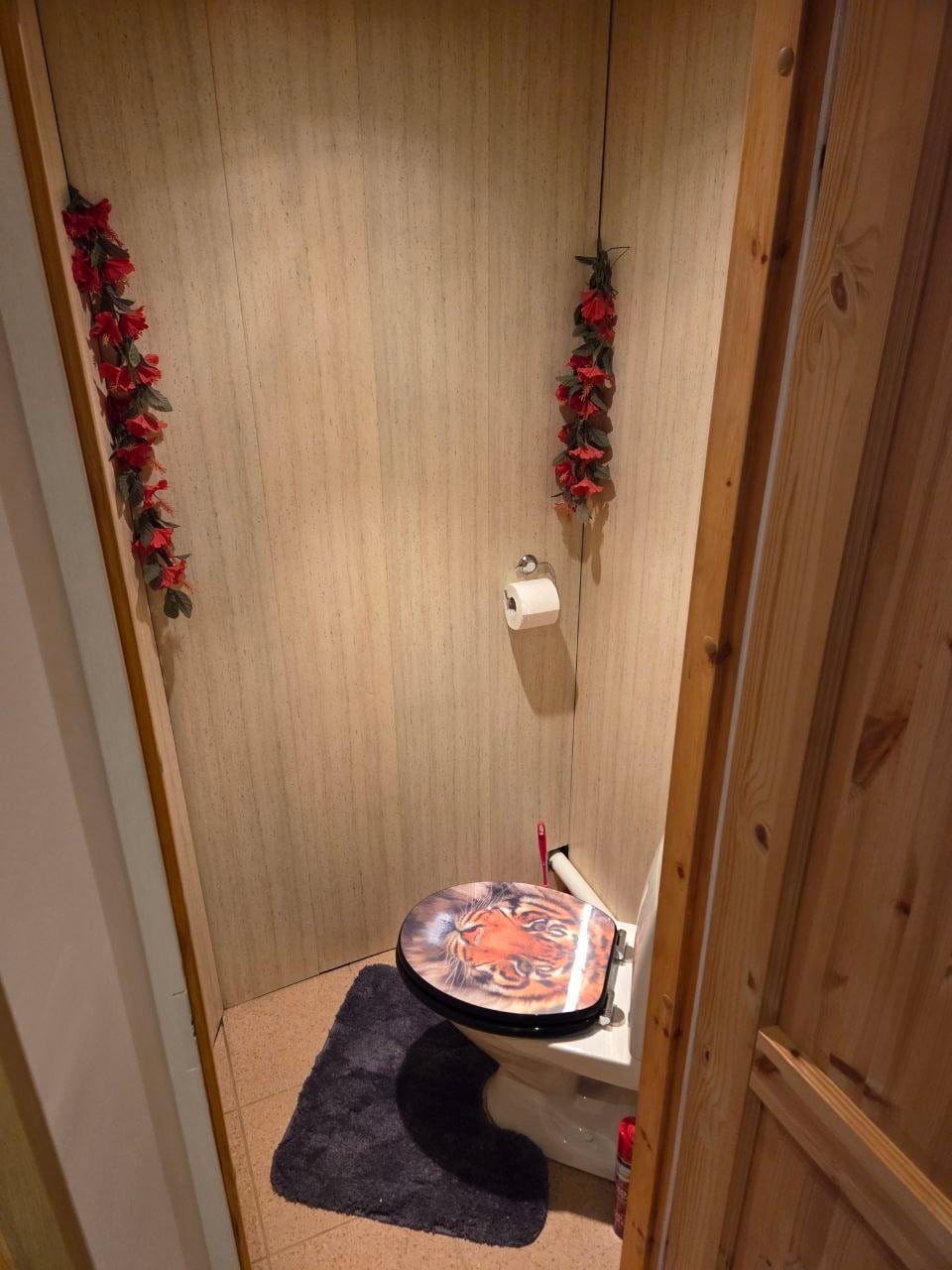 A small, private bathroom features a toilet adorned with a decorative seat cover. The walls are lined with textured wallpaper, while artificial flowers hang beside the toilet, adding a touch of color. A dark rug provides a soft surface underfoot.