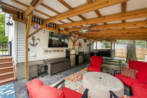 An outdoor patio offers a shaded space with a wooden canopy. Comfortable red chairs surround a round coffee table, complemented by a patterned rug. A large television is mounted on the wall above a grill and storage area, enhancing the inviting gathering spot.
