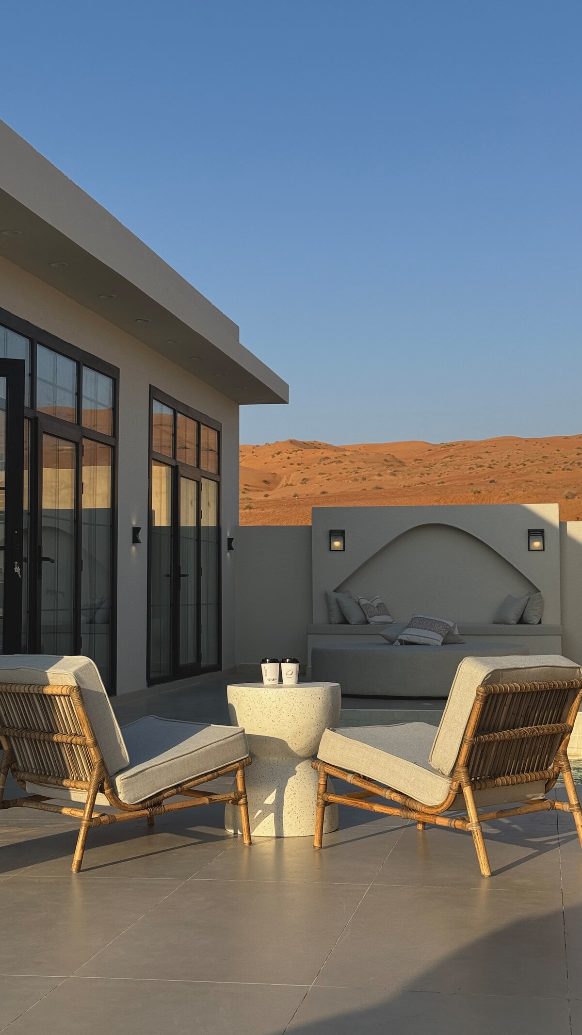 An outdoor seating area is featured, consisting of two rattan chairs beside a stone table. A cozy nook with soft cushions is positioned against a backdrop of sandy-colored hills. Large glass doors offer views of both spaces under a clear blue sky.