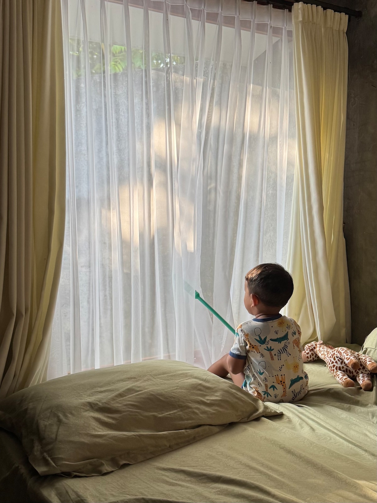 A child sits on a spacious bed, gazing out at sheer white curtains that softly diffuse natural light. Plush bedding in neutral tones provides a relaxed ambiance. A small stuffed animal rests beside the child, enhancing the cozy setting.