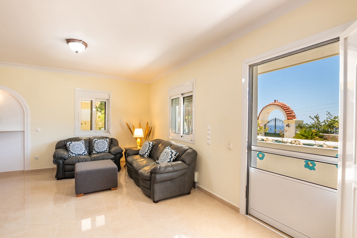 A living area features two comfortable leather sofas with decorative cushions, positioned around a small ottoman. Natural light enters through large windows, offering a view of the outdoors. Light-colored walls and tiled flooring create a bright and spacious environment.