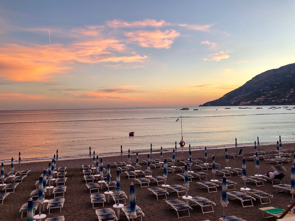 A serene beach scene is captured during sunset, with numerous lounge chairs arranged in neat rows on the sandy shore. The calm sea reflects the soft colors of the sunset sky, while distant boats can be seen on the horizon and hills line the background.