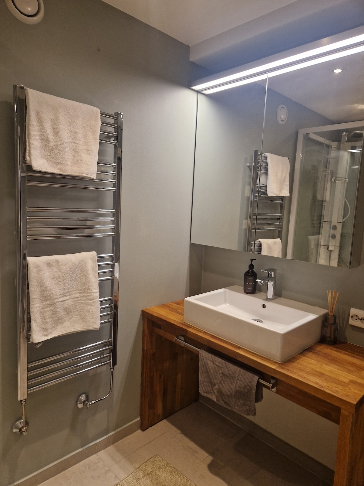 A modern bathroom features a sleek wall-mounted mirror above a white sink. A wooden vanity adds warmth, while a chrome towel warmer is mounted on the wall. Complementary tones create a clean and organized appearance, enhancing the overall functionality of the space.
