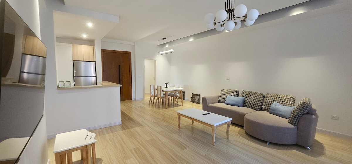 An open living space is shown, featuring a modern sectional sofa with soft cushions and a light coffee table. A dining area with four wooden chairs is seen in the background. The kitchen is equipped with stainless steel appliances, and warm light illuminates the room.