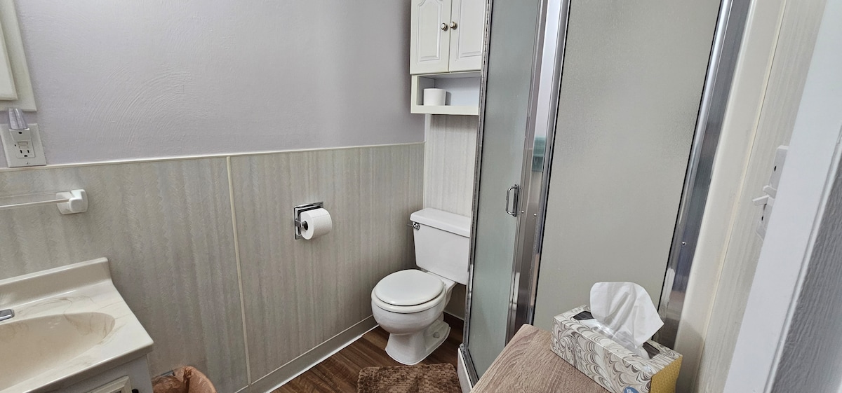 The bathroom features a light-colored vanity with storage above, a toilet, and a glass shower enclosure. Neutral tones are used throughout, with textured wall panels and a wooden floor. Basic amenities like tissue and toiletries are thoughtfully provided.