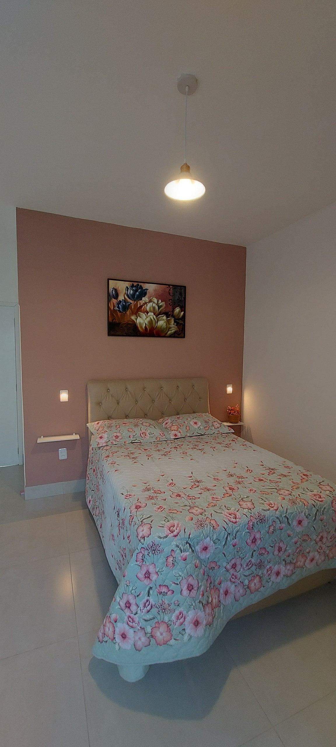 A bedroom is presented featuring a spacious bed adorned with a floral-patterned cover. Soft lighting is provided by wall-mounted fixtures on either side of the bed, while a vibrant piece of artwork is displayed above the headboard, contributing to the room's warm ambiance.