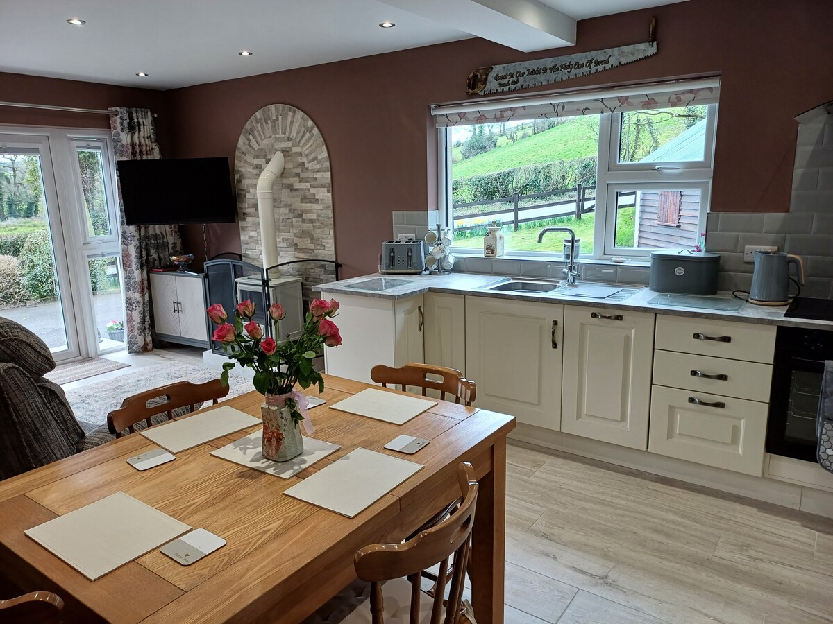 A well-appointed kitchen and dining area features a wooden table with six chairs, illuminated by natural light from large windows. Modern appliances are arranged on the countertop, complemented by decorative elements such as a vase of fresh flowers. A cozy seating area is visible in the background.