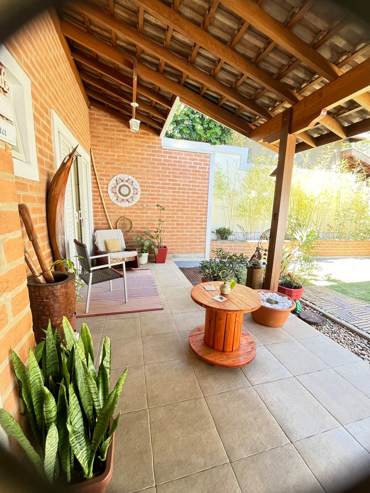 The cozy porch area features a wooden table surrounded by potted plants and a comfortable chair. A textured mat decorates the floor, while a brick wall and an overhanging roof provide shelter. Natural light illuminates the space, enhancing the connection with the outdoors.