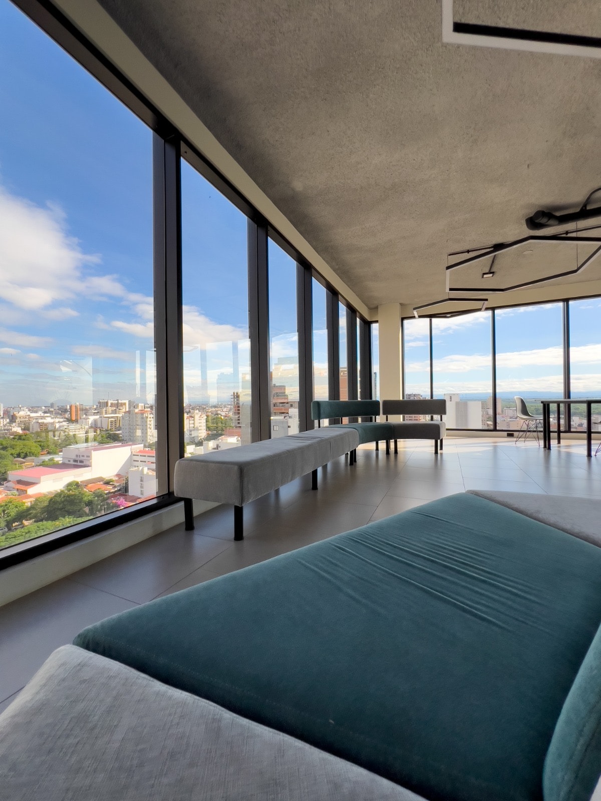 A spacious living area is defined by large floor-to-ceiling windows, offering expansive city views. Sleek benches are positioned along the windowsills, complemented by a comfortable sofa in muted tones. A minimalist light fixture hangs from the ceiling, enhancing the modern design.