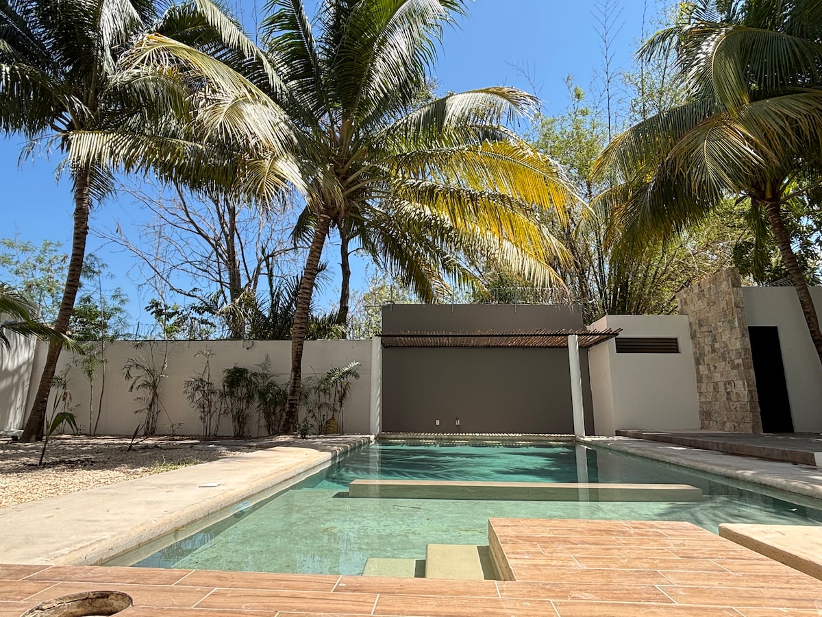 A serene swimming pool is surrounded by lush palm trees, providing a refreshing atmosphere. The pool features a shallow area and is complemented by stone-like tiles along the edge. A wall and plants are visible in the background, adding to the tranquil setting.