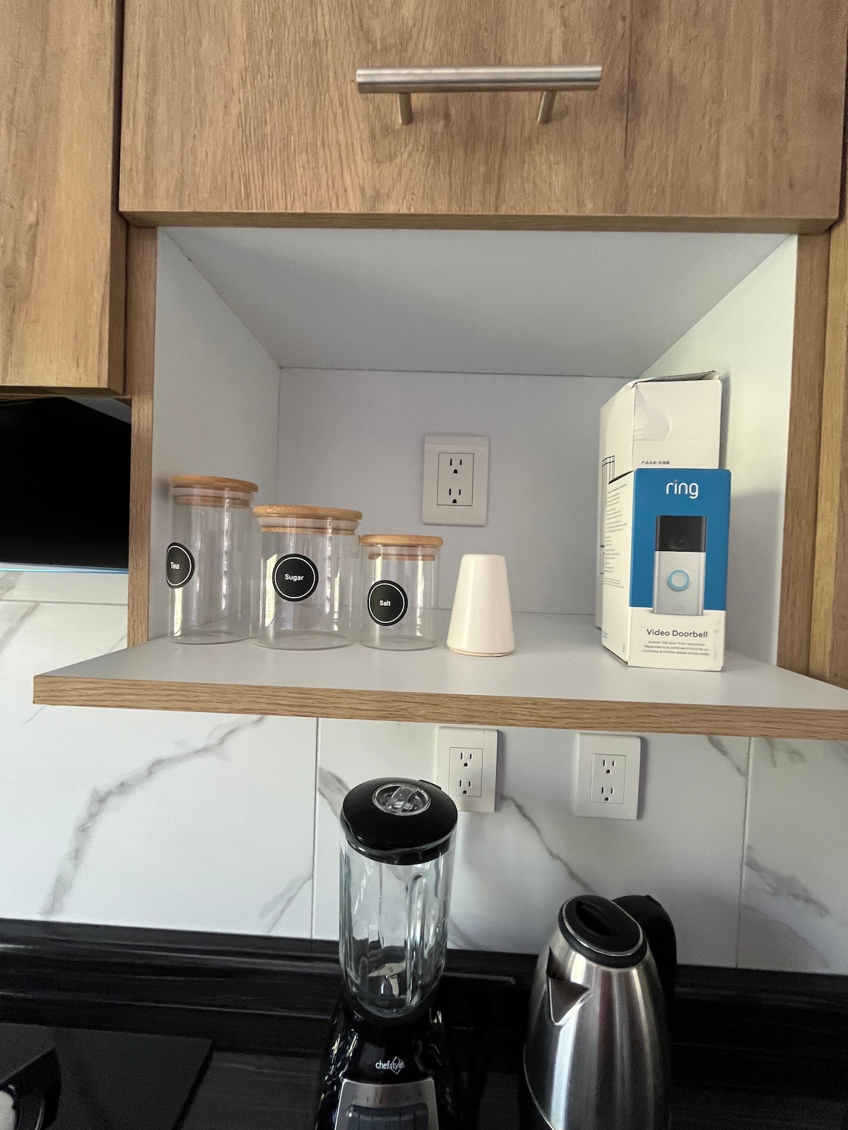 A modern kitchen shelf showcases four glass storage containers with wooden lids, offering a minimalist aesthetic. A small white object is positioned beside the containers. An electronic device box is visible, alongside a blender and a kettle on the countertop beneath.