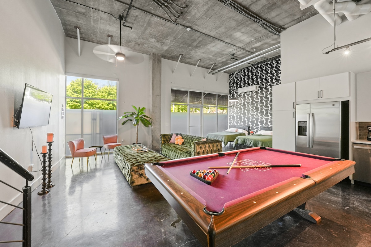 An open living area features a vibrant pool table with a set of colored balls, complemented by a spacious sofa and two modern chairs. Large windows allow natural light to fill the space, presenting a view of lush greenery outside. Decorative elements throughout the loft reflect a stylish design.