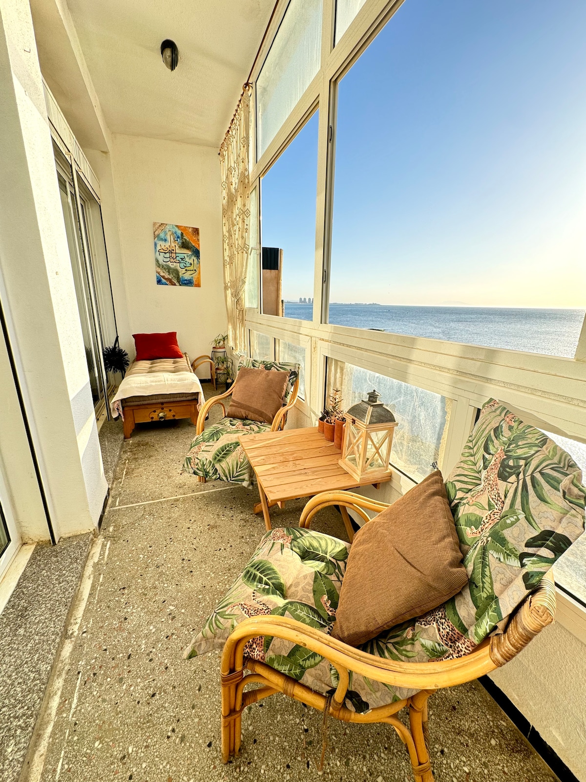 A sunlit balcony features two rattan chairs adorned with leafy-patterned cushions, creating a relaxed seating area. A small wooden table holds a lantern, while a daybed invites leisure with a soft throw. Expansive windows offer views of the ocean and sky beyond.
