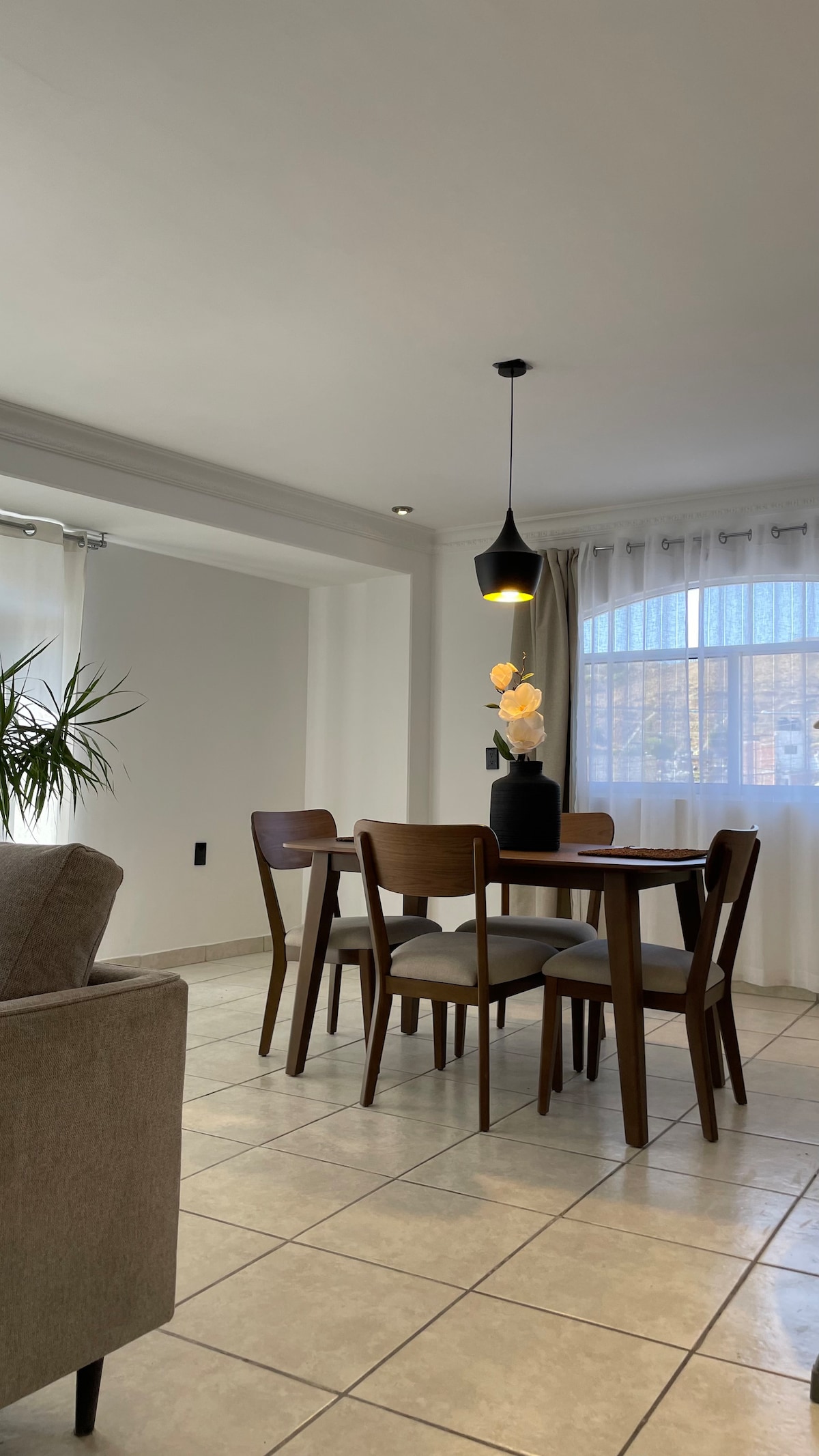 A dining area features a wooden table surrounded by four chairs. Natural light filters through sheer curtains near a large window. A modern pendant light hangs above the table, while a decorative vase with flowers sits as a centerpiece.