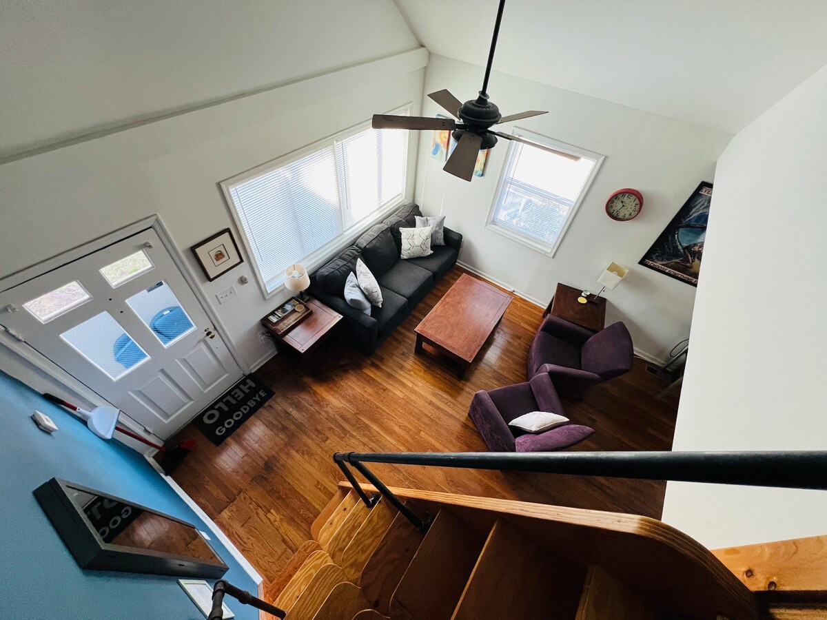 An aerial view captures the inviting living area, featuring a dark sectional sofa paired with a wooden coffee table. A cozy armchair is positioned near a bright window, allowing natural light to fill the space. Stairs are visible, leading to an upper loft area.