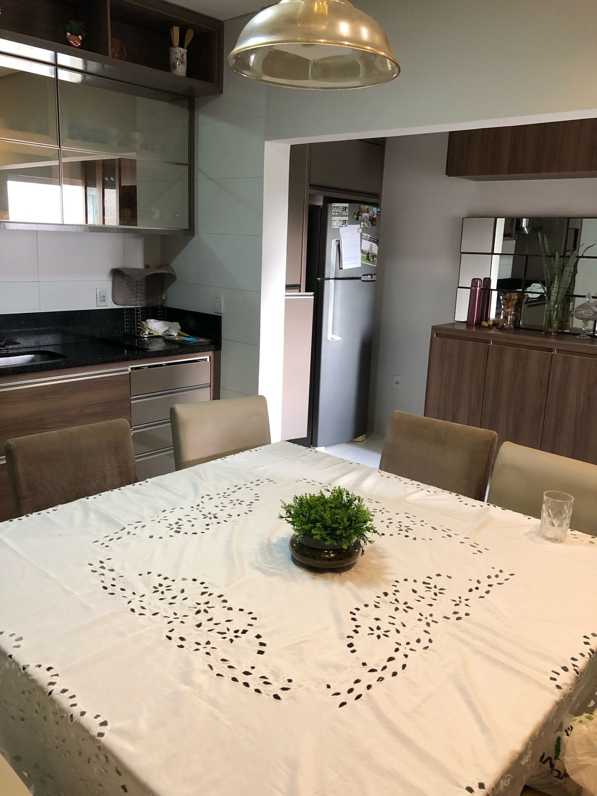 A dining area features a simple table set with a decorative cloth and a small potted plant at its center. Surrounding the table are four light-colored chairs. The open kitchen is visible in the background, showcasing modern cabinetry and appliances.