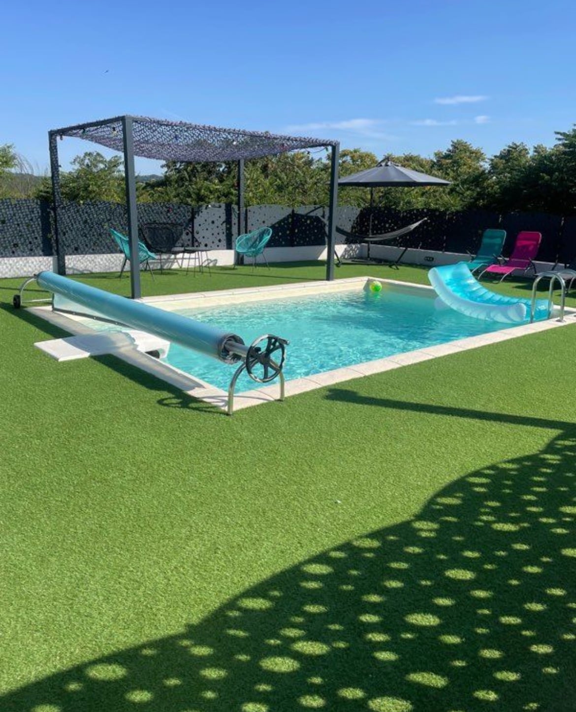 An outdoor swimming pool is featured, surrounded by artificial grass. A slide extends into the water, and two colorful lounge chairs are positioned nearby. A shaded area with a canopy offers a respite from the sun, creating a relaxing atmosphere.