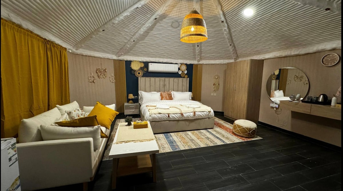 The room features a spacious bed with a textured headboard, accompanied by cozy seating on a stylish sofa. A neutral colour palette is complemented by warm yellow curtains. Decorative elements include woven wall art and a round mirror, with a compact table and kitchen area visible.