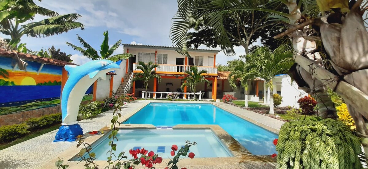 The image showcases a large pool surrounded by vibrant landscaping, featuring lush palm trees and colorful flowers. A playful dolphin statue is positioned near the pool's edge. A two-story building is visible in the background, with patio seating under an overhang.