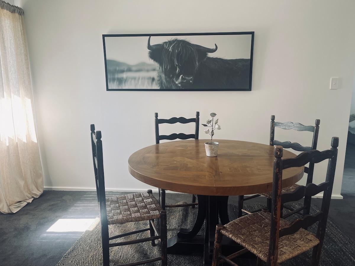 A round wooden dining table is surrounded by four rustic chairs, two of which feature woven seats. A monochromatic artwork of a Highland cow is displayed on the wall above the table. Soft natural light filters in through sheer curtains, enhancing the warm atmosphere.