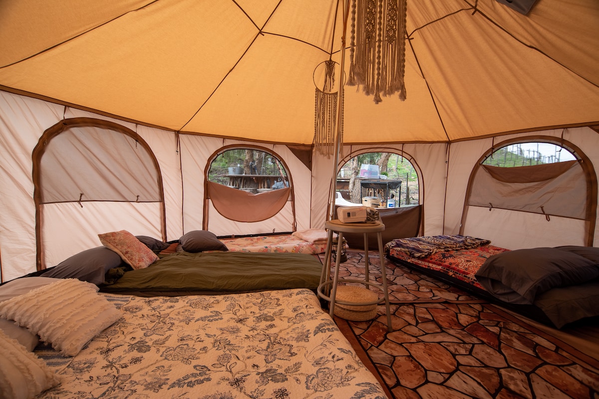 A spacious glamping tent interior features several comfortable beds arranged on a textured floor. Soft bedding and decorative quilts are visible. Natural light enters through multiple large windows, enhancing the cozy ambiance. A small table is placed centrally, providing a spot for decor and personal items.