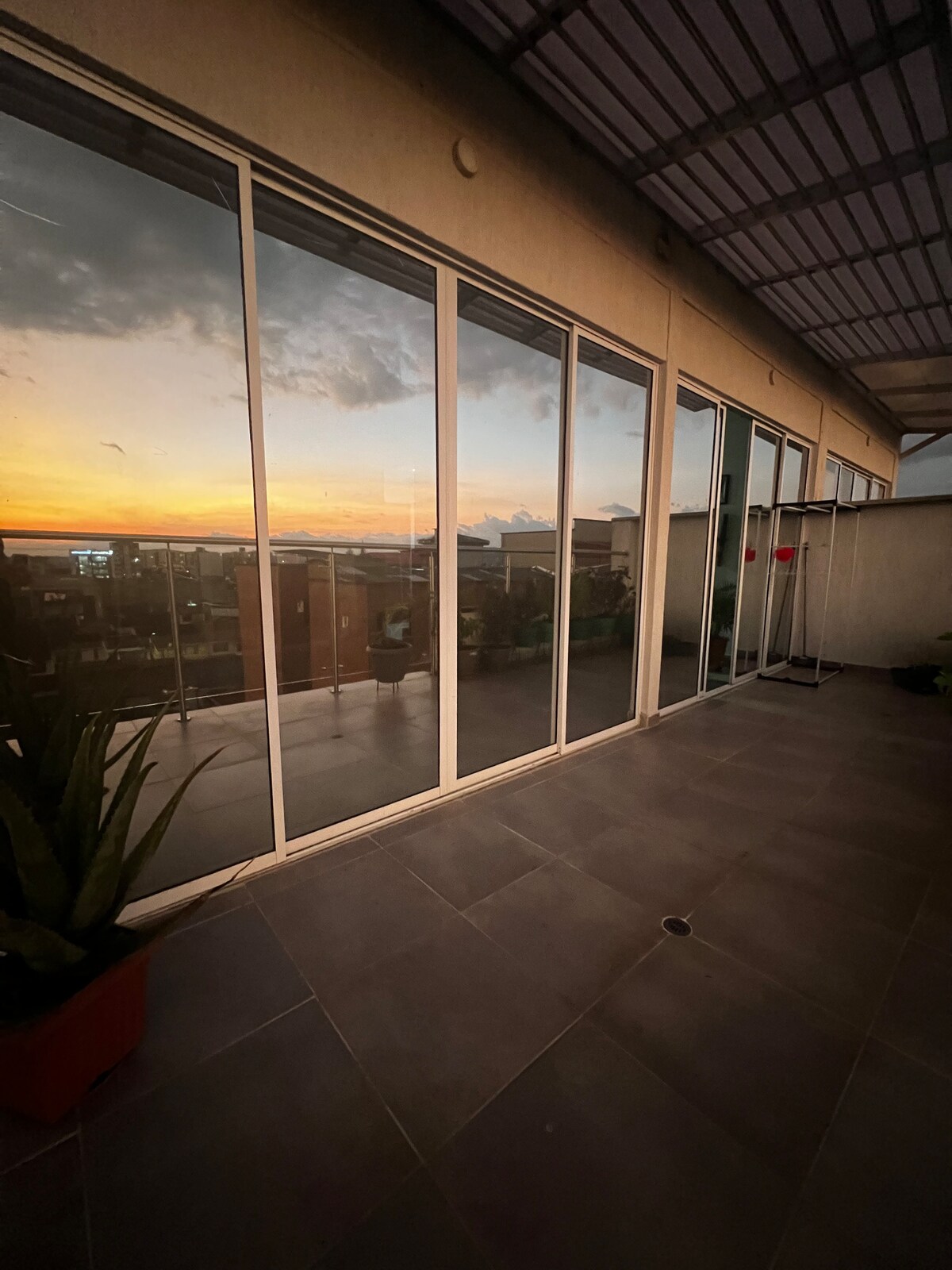 A spacious terrace is visible, marked by large glass doors reflecting a colorful sunset. Potted plants are seen along the edge of the terrace. The tiled floor and open space invite comfortable outdoor relaxation, while additional elements hint at a well-designed living space.