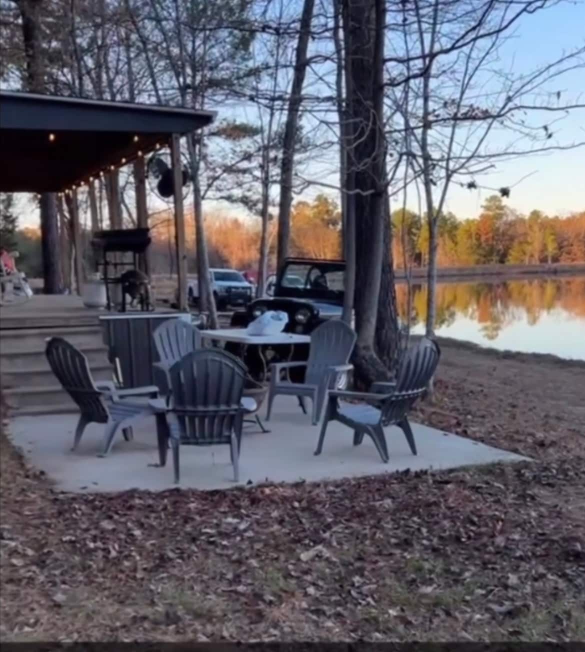 A patio area is situated near a tranquil lake, surrounded by trees. Several Adirondack chairs are arranged around a circular table, providing a space for relaxation. Soft evening light reflects off the water, creating a peaceful environment.