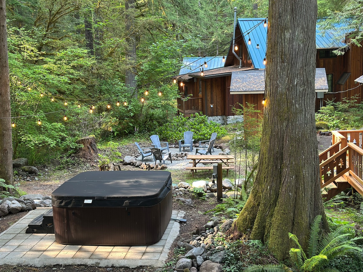 A hot tub is positioned on a paved area surrounded by lush greenery. Soft lights hang above a seating arrangement featuring several chairs and a table, creating a cozy atmosphere. A wooden pathway leads to the cabins visible in the background.