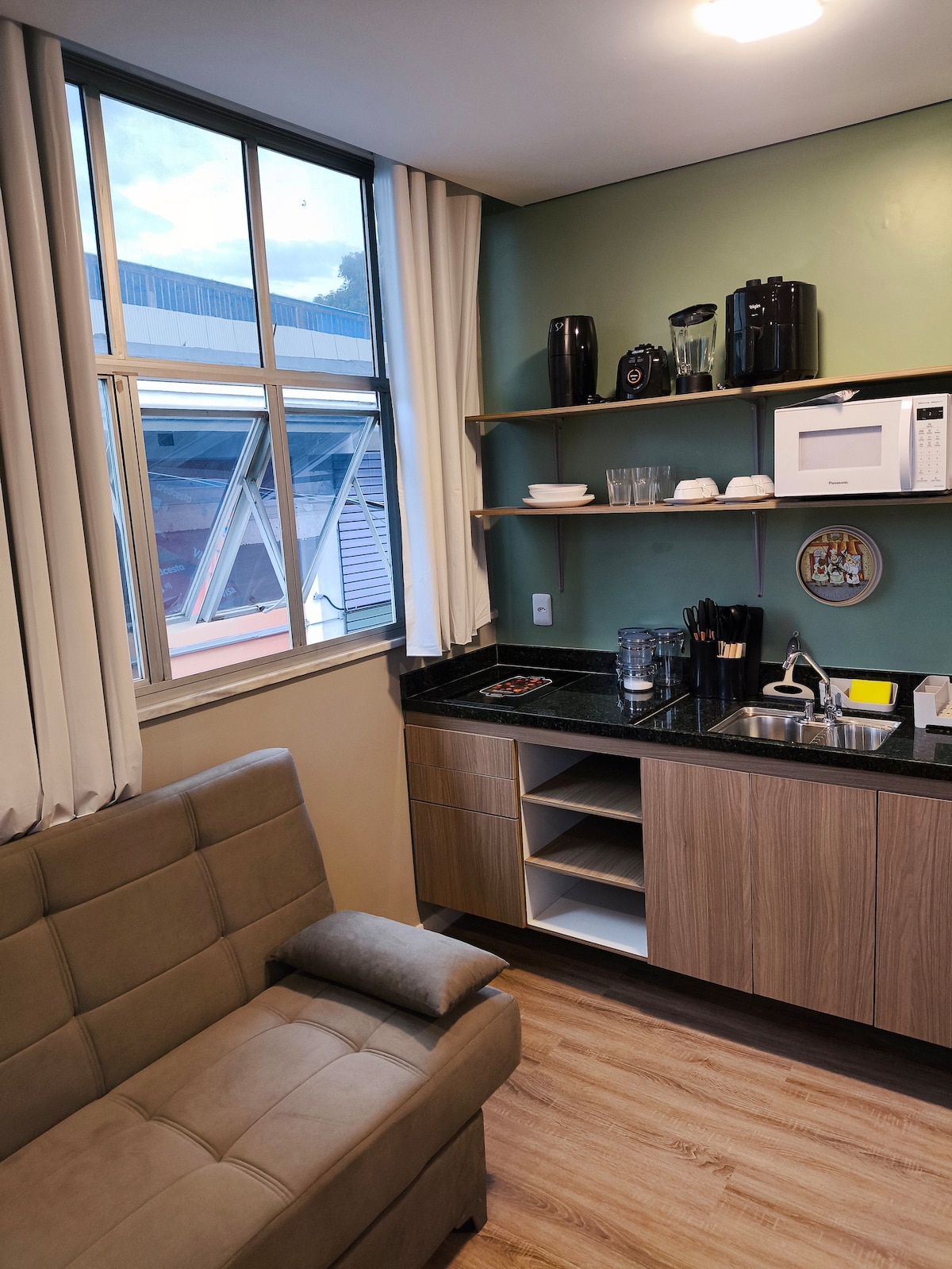 The kitchenette is equipped with modern appliances including a microwave and coffee maker. A small dining area features a sleek countertop, while a cozy sofa adds comfort. Natural light filters through the window, highlighting the organized shelving with glassware and utensils.